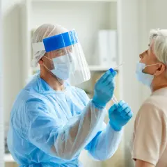 Arzt in Schutzkleidung nimmt bei älterer Frau einen Rachenabstrich. Testet man so auch auf MRSA, was ist das?