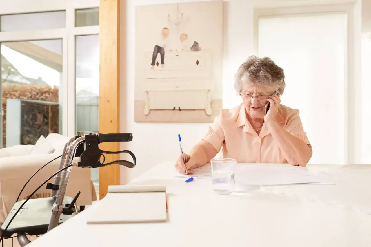 Ältere Dame mit Pflegegrad 2 telefoniert und schreibt, ihr Gehwagen steht bereit.